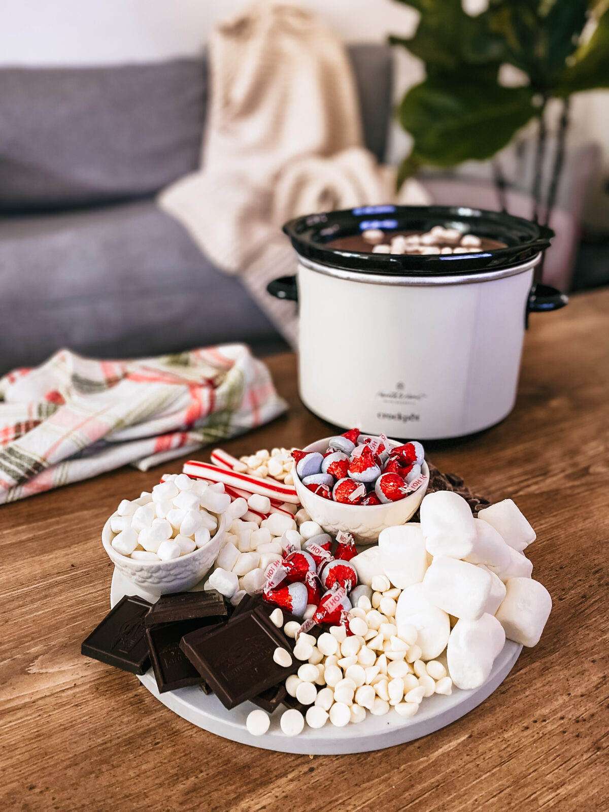 A hot cocoa charcuterie board with marshmallows, hershey kisses, chocolate chips, and peppermint sticks on a table with a crockpot filled with hot chocolate.