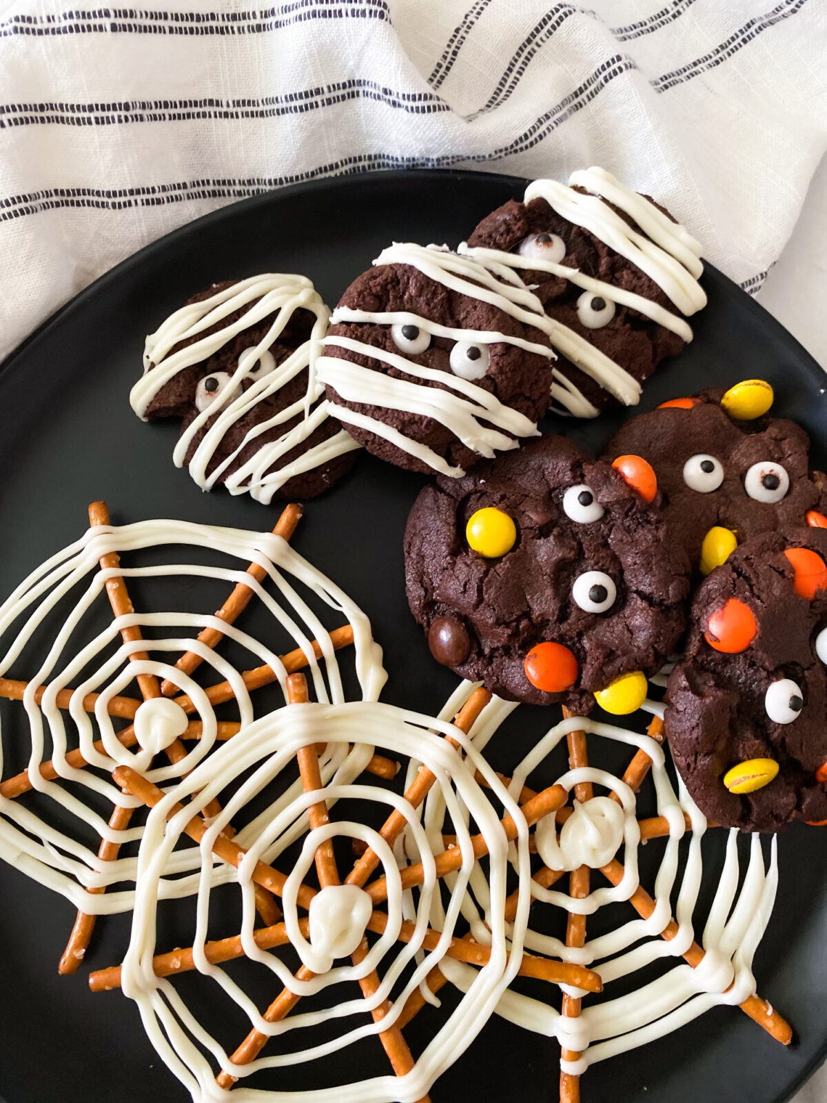 Halloween treats plate with spider web pretzels, mummy cookies, and m&m cookies. 