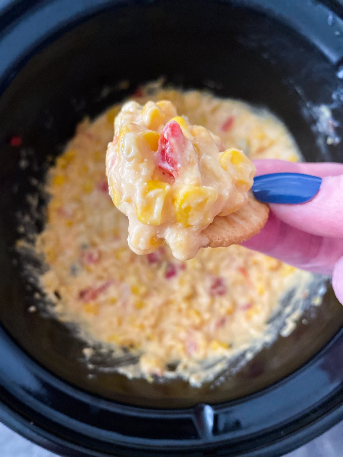 A woman's hand holding a cracker over a Crockpot with a scoop of Corn Dip.