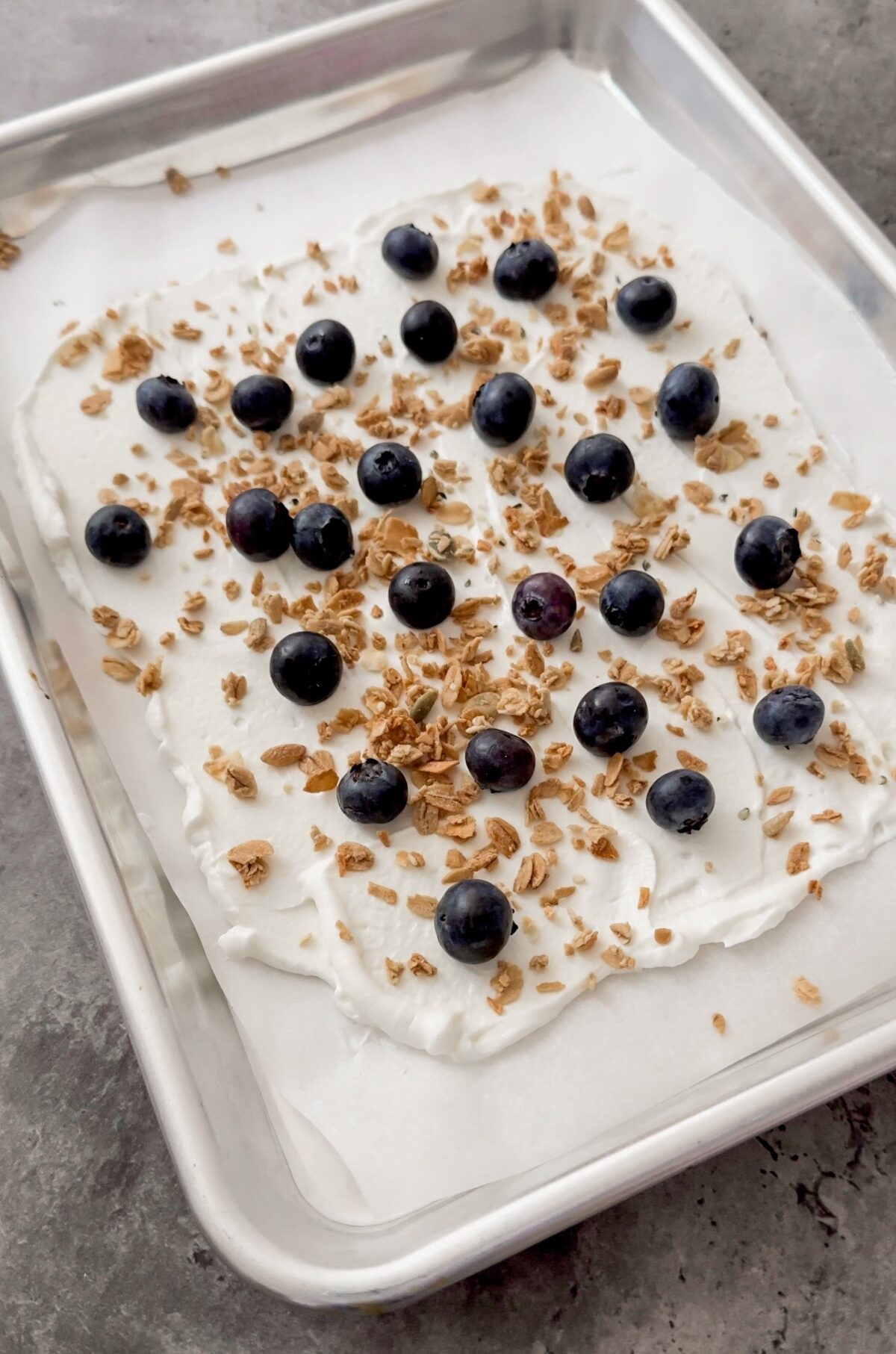 blueberries and granolas sprinkled on a layer of yogurt spread on cookie sheet
